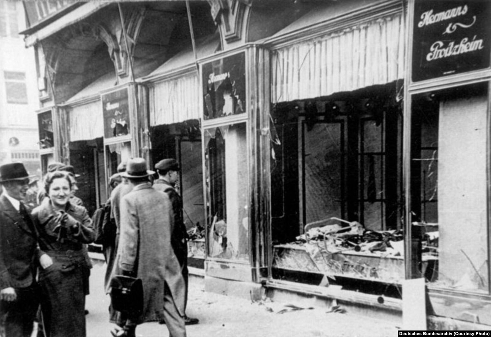 A row of shops in Magdeburg, Germany, the morning after Kristallnacht, with plate-glass windows smashed and merchandise scattered. Passersby in coats and hats walk past; one woman is smiling.