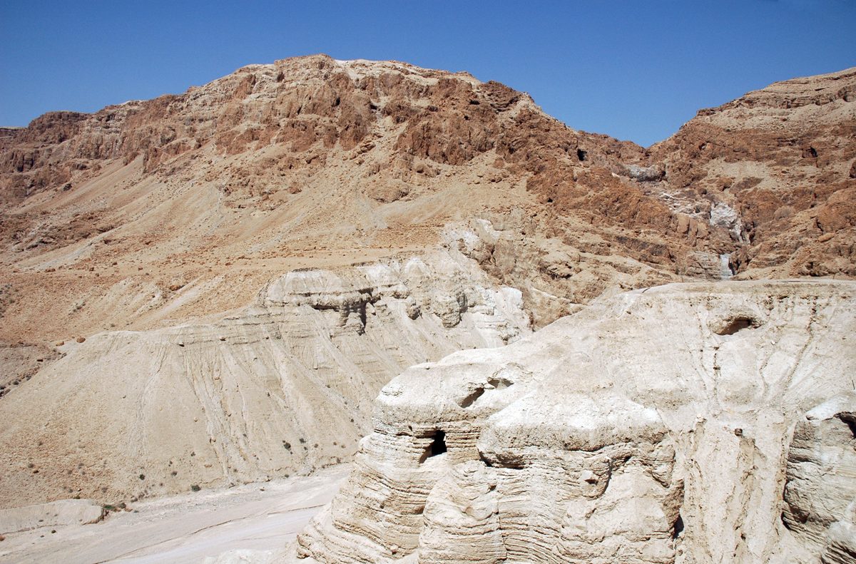 A pale, eroded desert cliff face riddled with dark cave openings, under a bright sky, above barren rocky ground.