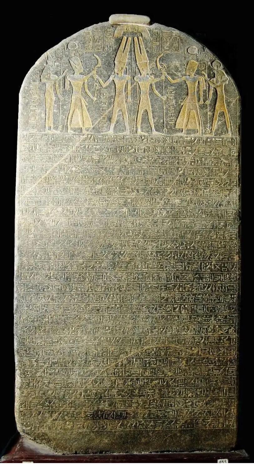 A tall dark-granite Egyptian victory stele, its rounded top carved with a pharaoh facing a god, and dense rows of hieroglyphs below.