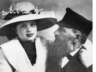 A studio portrait of a young woman in a wide-brimmed hat trimmed with flowers beside an older bearded man in a dark hat, from the People of a Thousand Towns collection documenting Jewish life in Eastern Europe