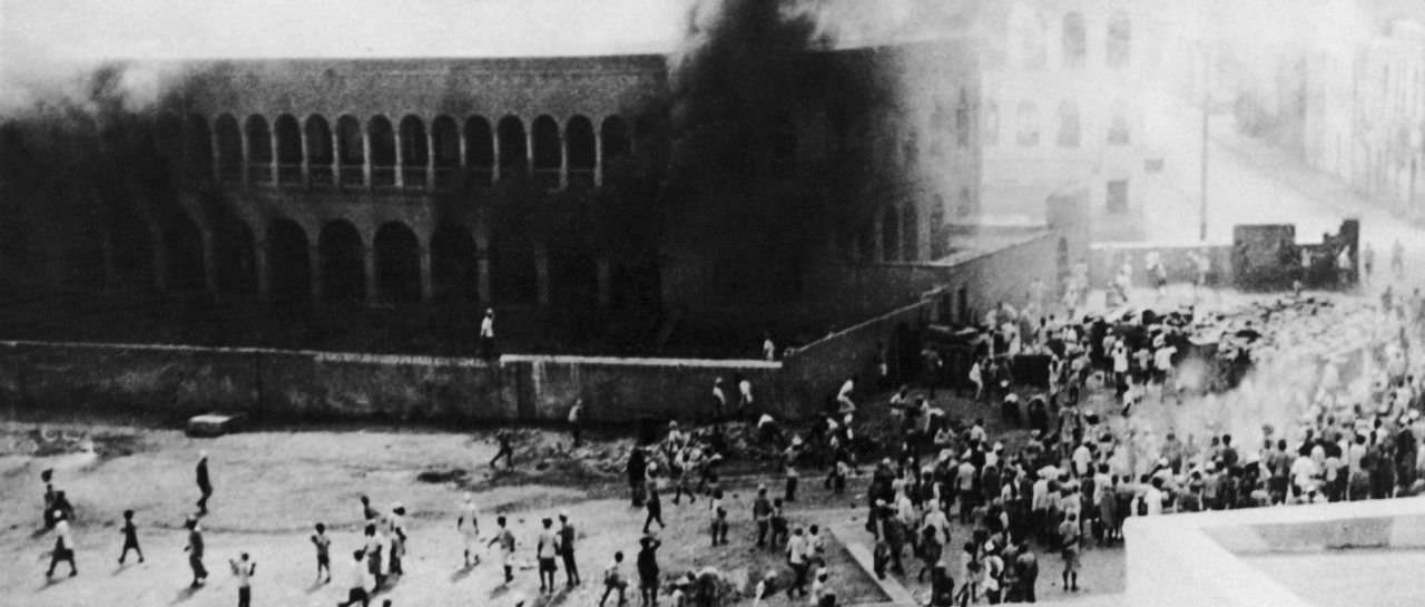 A large arcaded school building engulfed in black smoke during the December 1947 Aden pogrom, with crowds scattered across the open ground below.
