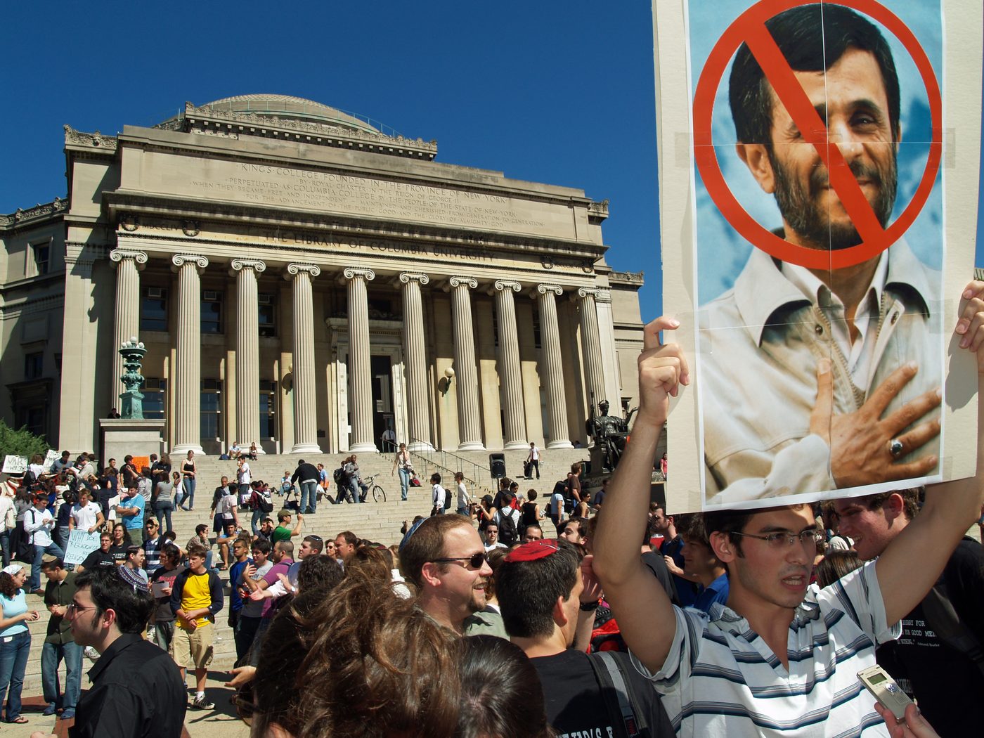 A dense crowd of protesters on Columbia’s campus in front of the Low Library steps, one holding a poster of Ahmadinejad crossed out in red.