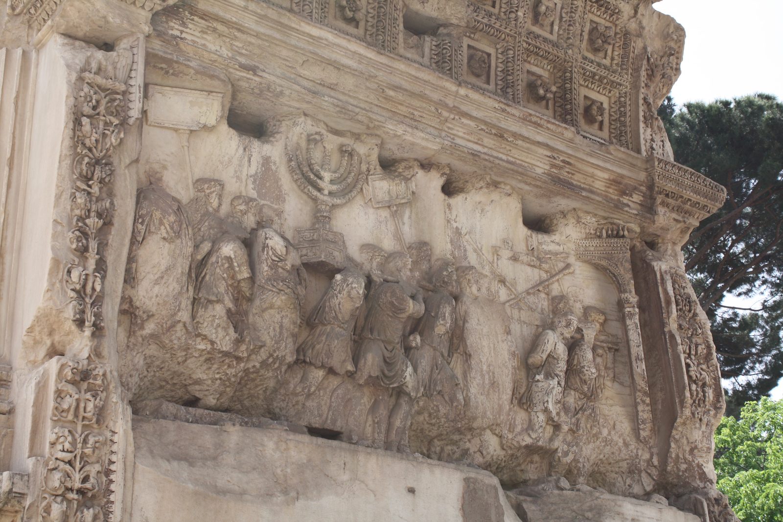 A weathered carved stone relief showing a crowd of figures carrying a large seven-branched lampstand and other treasures in a procession.