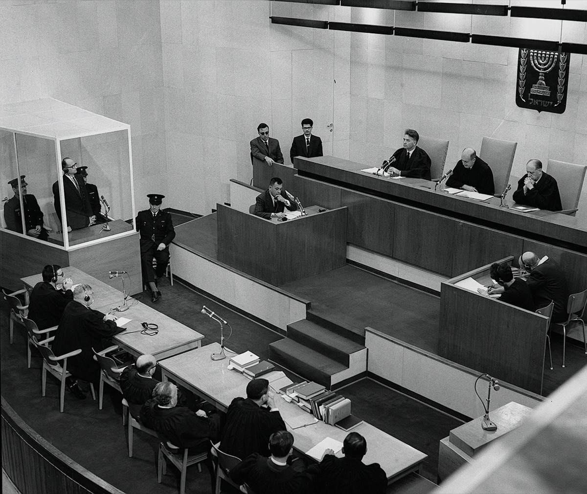 A wide view of the Eichmann trial courtroom in Jerusalem: Adolf Eichmann seated in a glass booth at left, guards beside him, judges on the bench beneath the emblem of Israel.