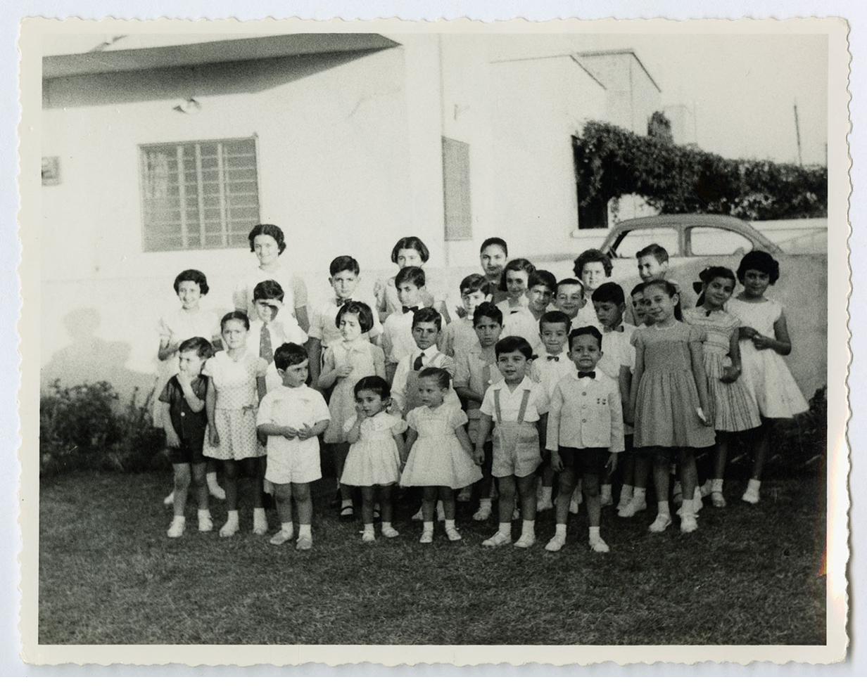 A group of about two dozen Jewish children in party clothes: boys in shorts and bow ties, girls in dresses, standing in a garden in Baghdad in 1959, with a few adults behind them and a 1950s car at the edge of the frame.