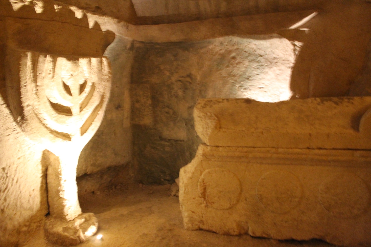 A large menorah carved into the limestone wall of an underground burial chamber, next to a plain stone sarcophagus.