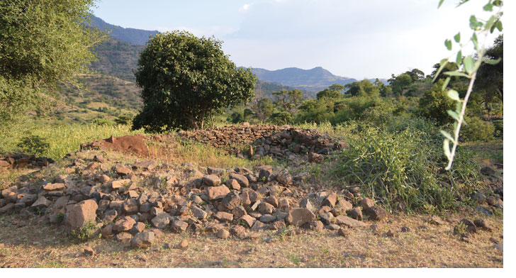 The stone foundations of a Beta Israel prayer house, now a ruin, set in the green Ethiopian highlands with mountains behind.