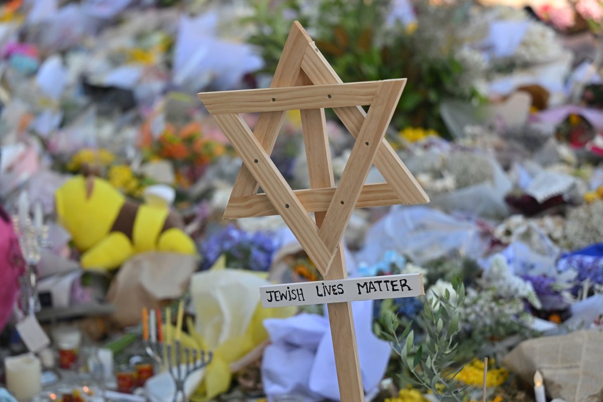 A large tribute of flowers, candles, and an Israeli flag covers the ground at the Bondi Pavilion in Sydney, with mourners and press gathered behind a barrier.