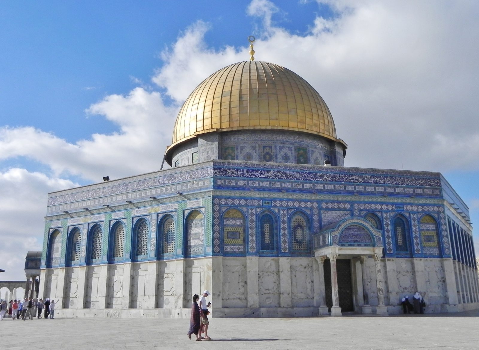 The Dome of the Rock under a blue sky: a gold dome on an octagonal building faced with blue, green, and white patterned tiles and bands of Arabic inscription, a few people crossing the stone plaza in front.