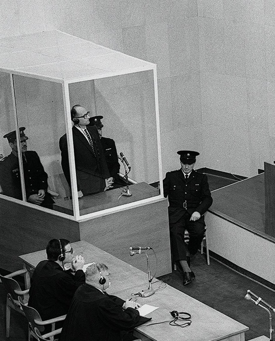 The glass booth at the Eichmann trial: a bulletproof glass box on a white metal frame, Eichmann standing inside it wearing headphones, Israeli guards beside the glass, a microphone mounted at its front.