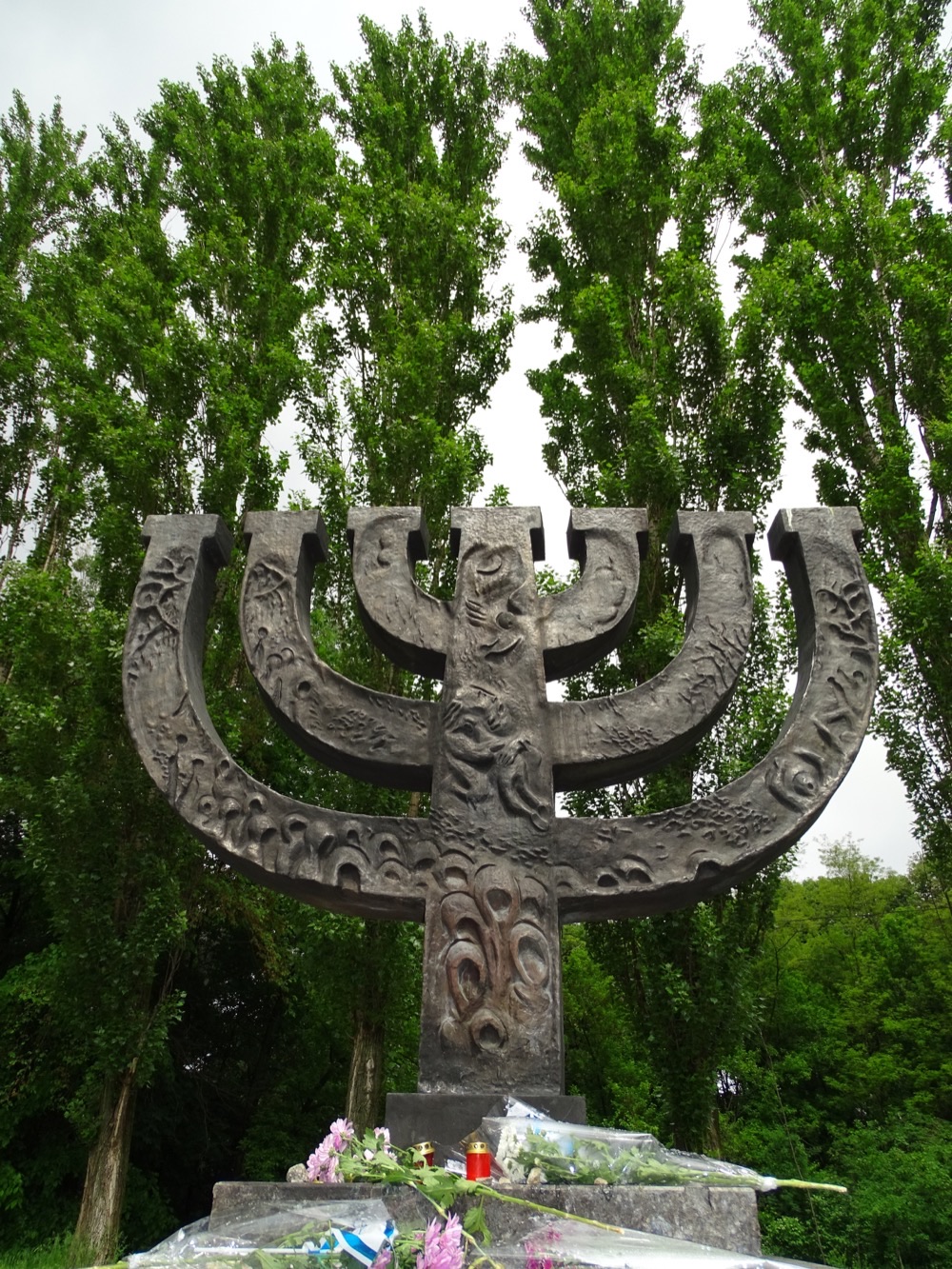 A large bronze menorah memorial sculpture among trees, with flowers and memorial candles at its base, at Babyn Yar.