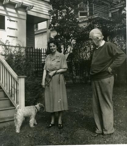 An elderly Einstein in a casual sweater stands in a yard beside a woman holding a small terrier on a leash, outside a wooden house.