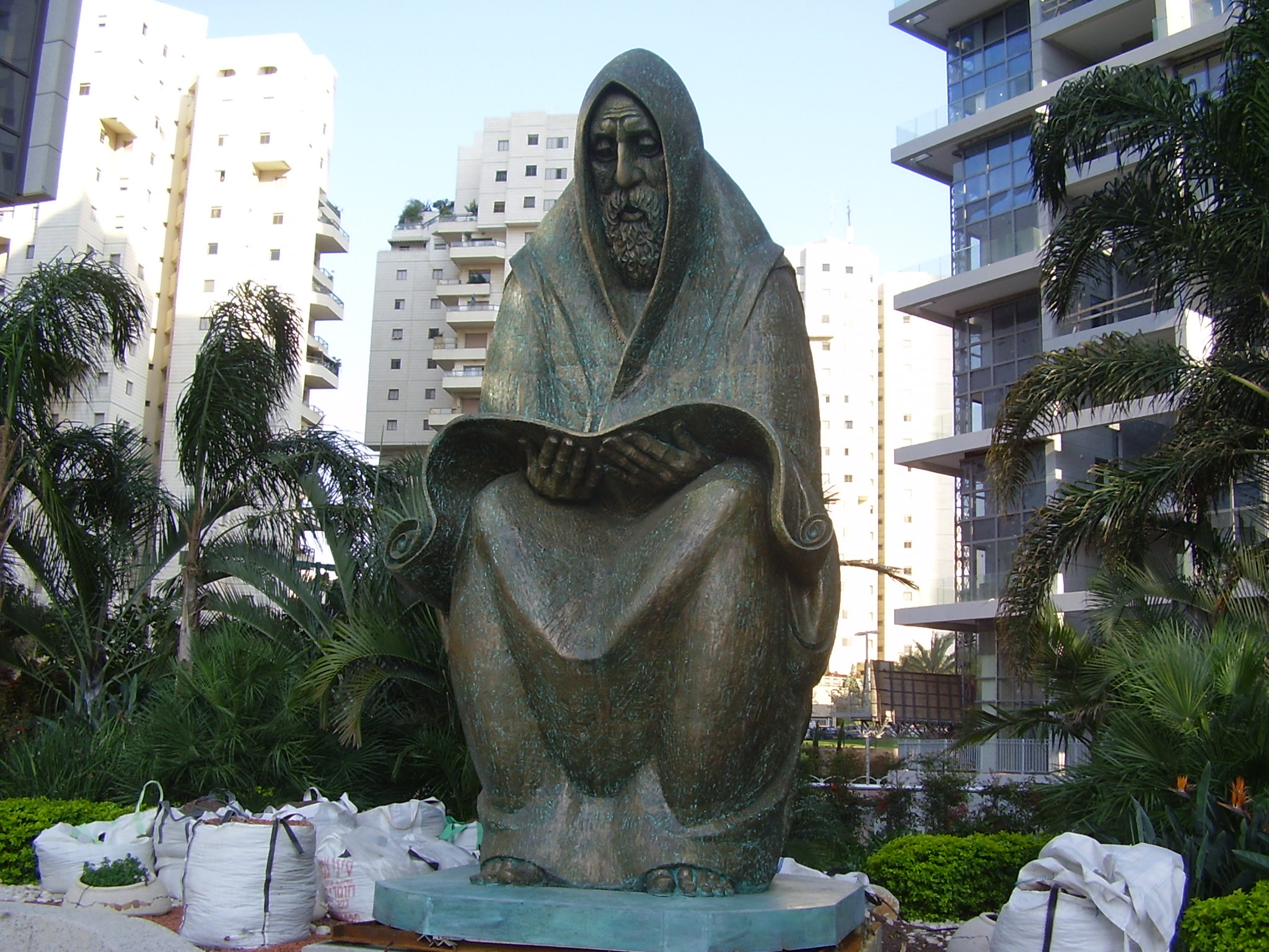 A weathered bronze sculpture of a seated, hooded figure bent over an open scroll, set on a stone base in a planted public square in Ramat Gan, with apartment buildings behind.