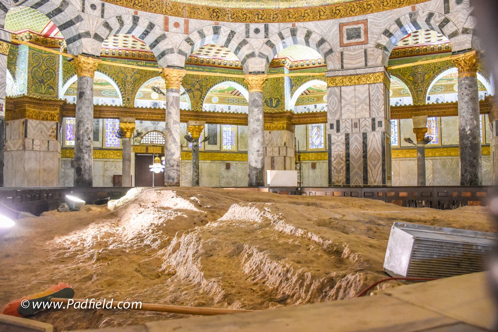 The interior of the Dome of the Rock: a ring of marble columns and striped arches under a richly decorated gold dome, surrounding a large mass of exposed pale bedrock at the floor's center.