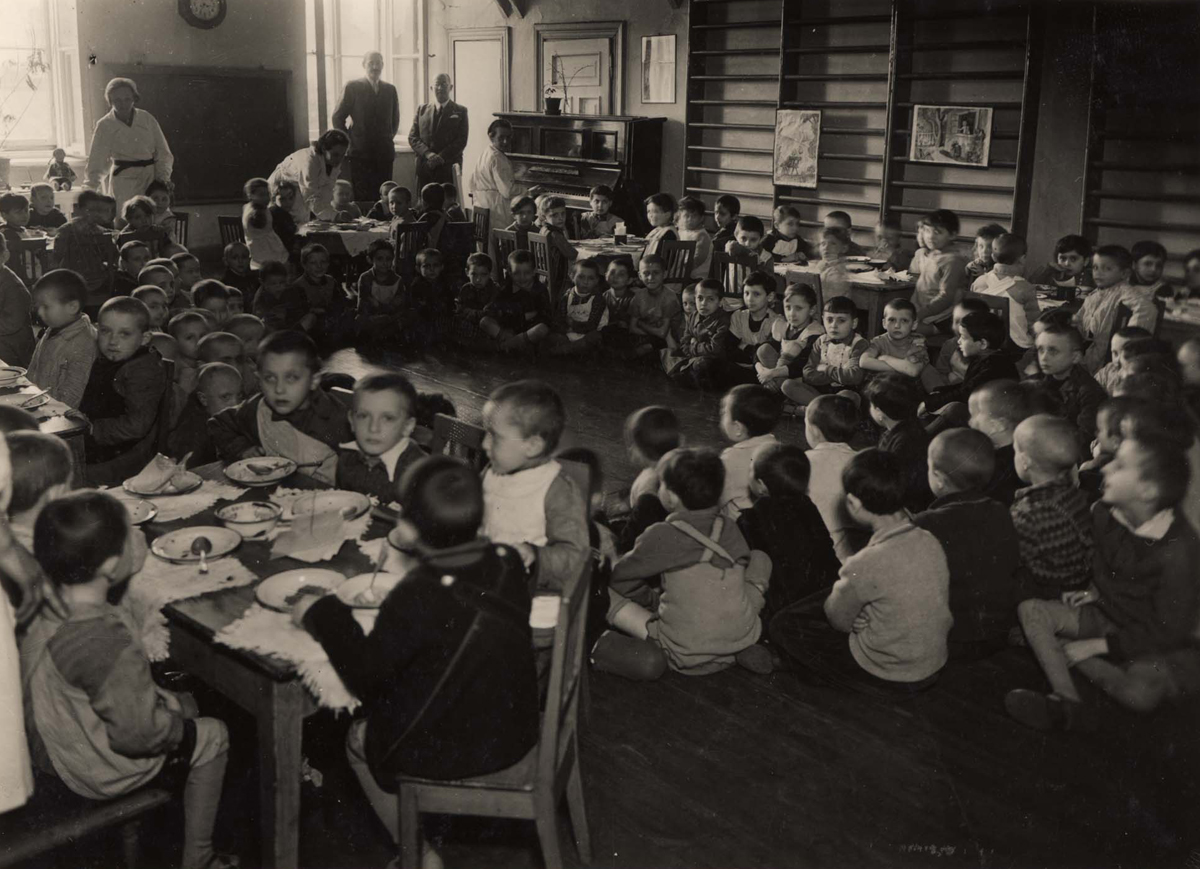 A crowded room of young children in the Warsaw ghetto, some seated at tables with bowls and plates, many more sitting on the floor, watched by a few adults near a piano and children's drawings on the wall.