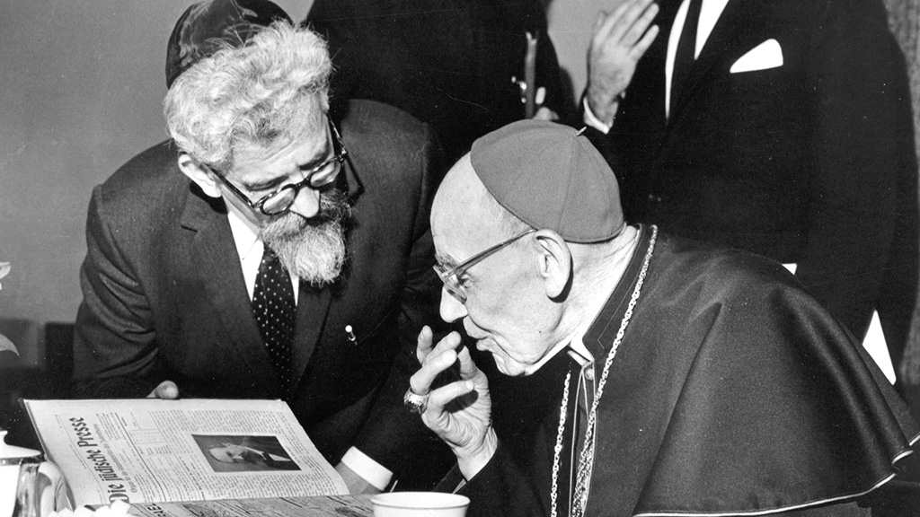 A black-and-white press photograph of Abraham Joshua Heschel, bearded and in a yarmulke, leaning over an open Jewish newspaper with Cardinal Augustin Bea, in skullcap and pectoral cross.