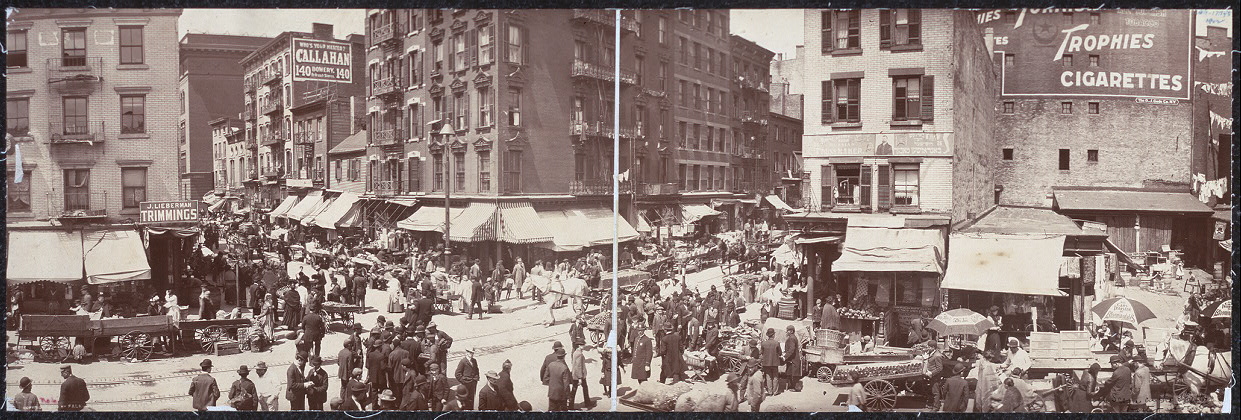 A panoramic photograph of Hester Street on the Lower East Side, crowded with pushcarts, awnings, shop signs, and people filling the street between tenement buildings.