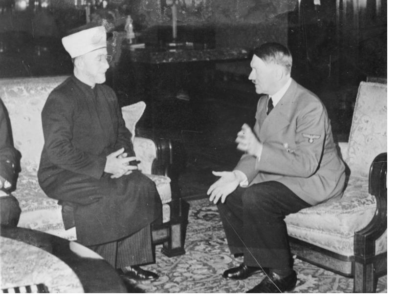 A black-and-white photograph of Hajj Amin al-Husseini, in clerical robes and turban, seated and speaking with Adolf Hitler, in uniform, in a furnished room.