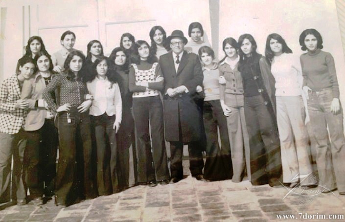 A black-and-white photograph of about fifteen teenage girls in 1970s clothing: flared trousers and patterned tops, gathered around an older man in a suit and hat, at a Jewish school in Iran.