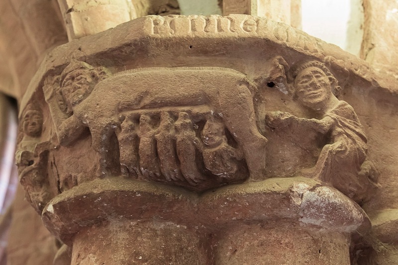 A carved sandstone column capital inside a church cloister: a sow in the center with small figures crouched beneath it and a human figure reaching toward it from the right.