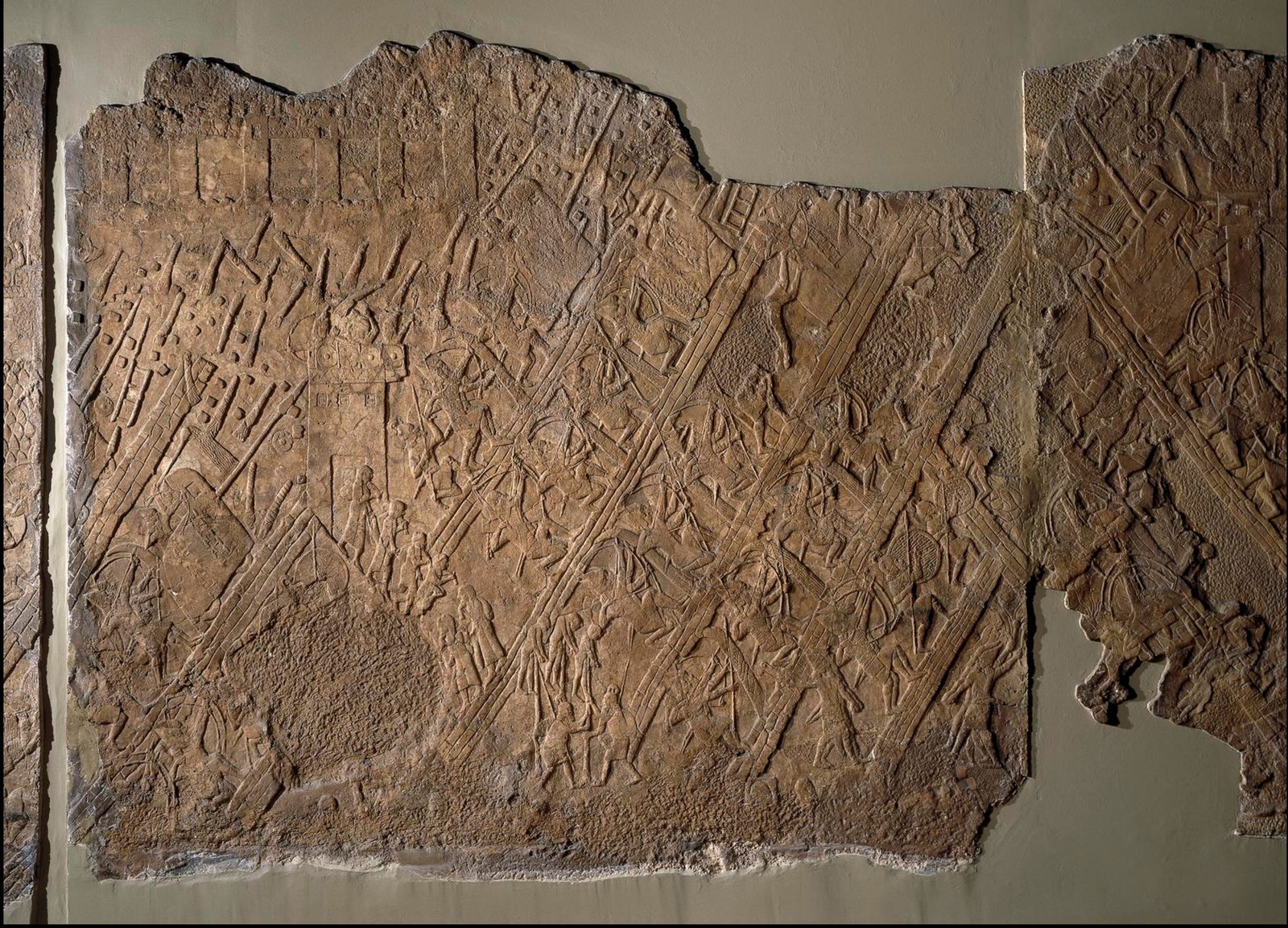 A long carved stone relief showing an Assyrian assault on a walled city: soldiers climb siege ramps, archers fire, and battering rams roll against the towers.