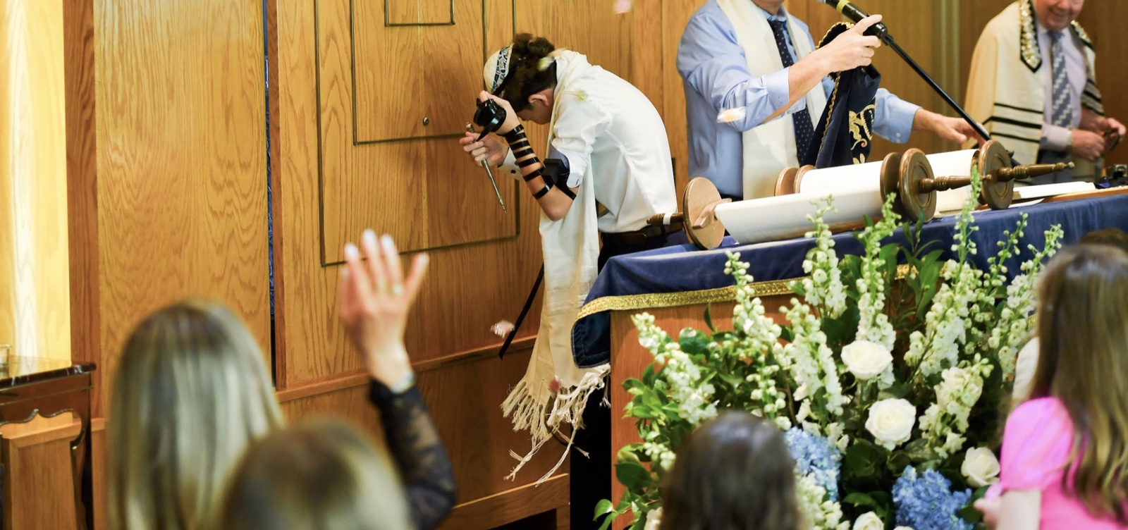 A bar mitzvah boy in tallit and tefillin bows beside the open Torah scroll as the congregation throws candy