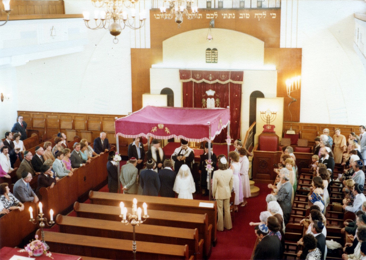A wedding ceremony under a canopy in a synagogue, the couple and rabbi beneath the chuppah, guests in the pews