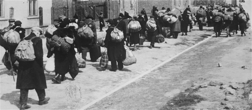 A long column of people walking down a Lodz ghetto street during deportation, each carrying bundles, sacks, and rucksacks on their backs, Star of David badges visible on their coats.