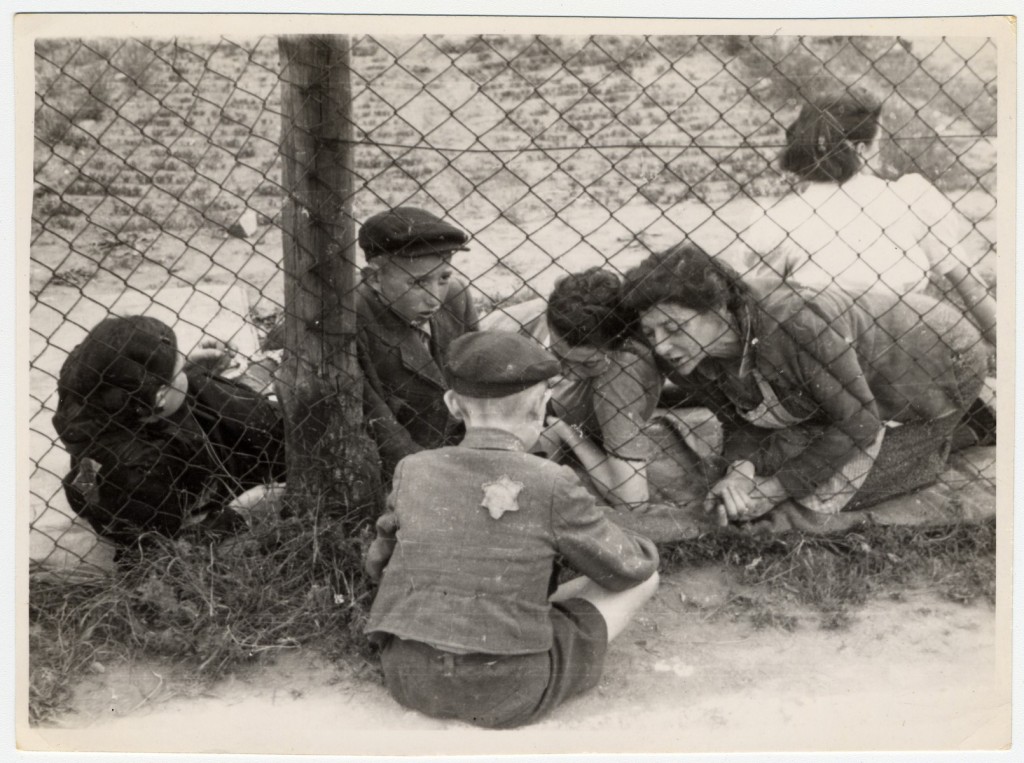 Through a chain-link fence, family members reach toward a child held on the other side; a boy with a Star of David badge sits against a post in the foreground.