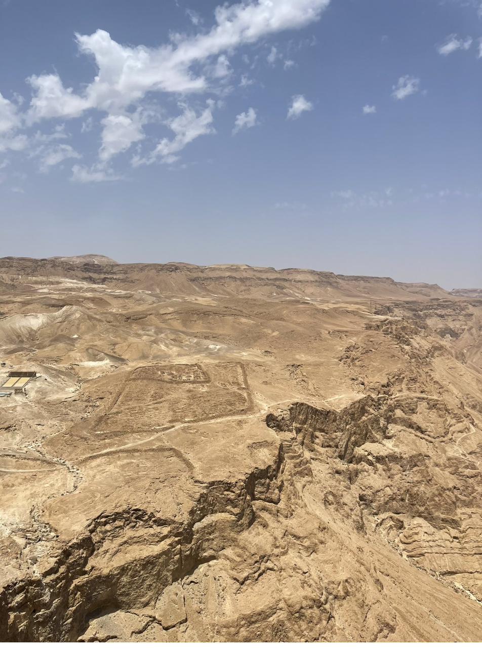 A view from a high desert plateau looking out over pale tan hills and a deep ravine, with faint rectangular outlines visible on the ground below.