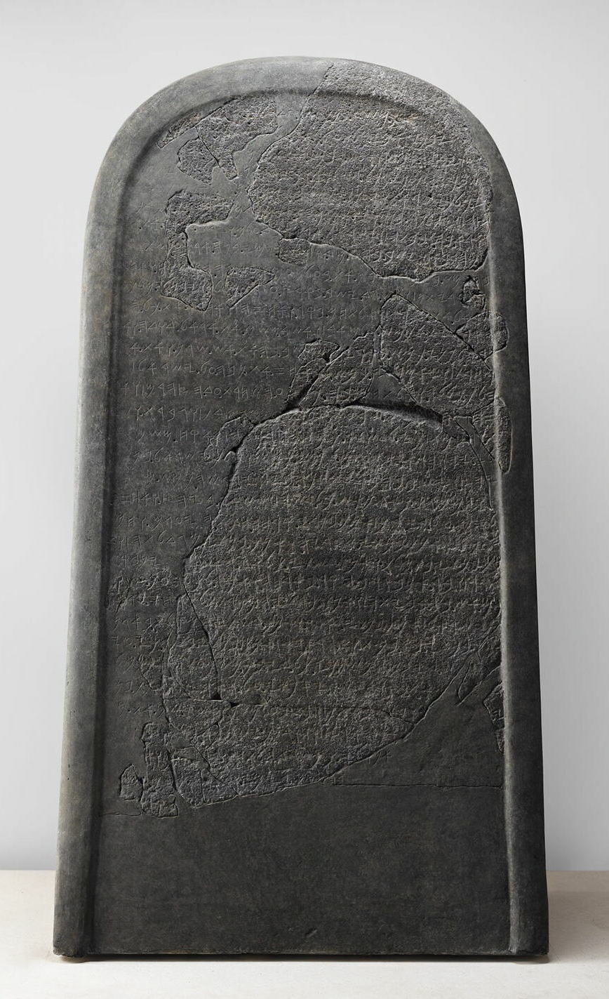 A tall dark basalt stele with a rounded top, its surface heavily cracked and patched, covered with rows of an early Semitic alphabet.