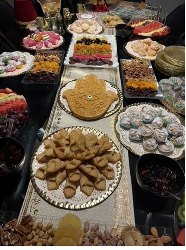 A festive table laden with North African Jewish sweets, pastries, dried fruit, and nuts for the Mimouna celebration, with a hamsa-shaped centerpiece.