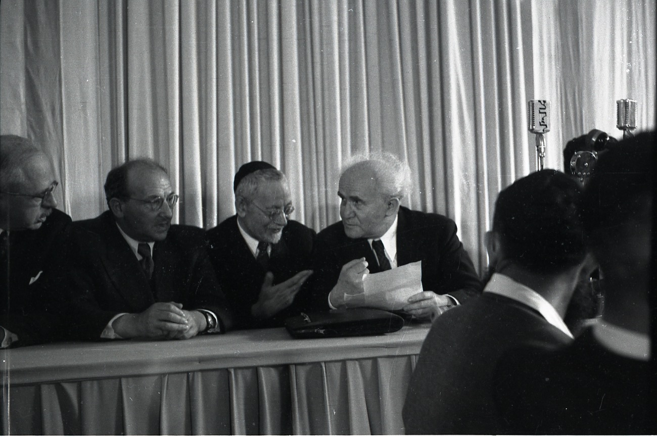 David Ben-Gurion seated at a table holding the declaration document at the founding ceremony, with other signatories and microphones, May 1948.