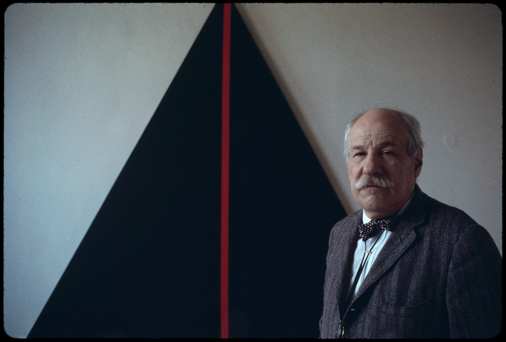 A 1969 color photograph of Barnett Newman in a tweed jacket and bow tie, standing beside a triangular black canvas split by a vertical red stripe