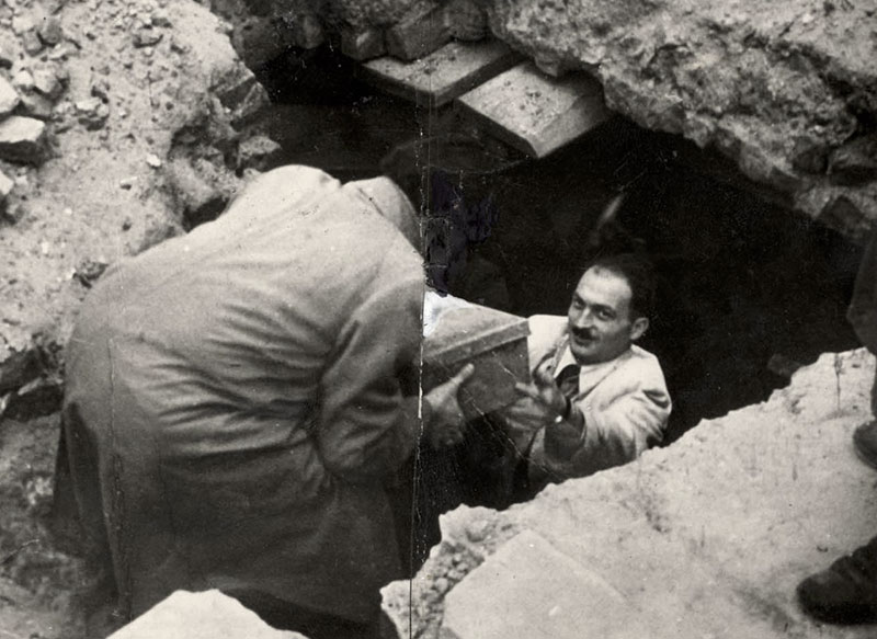 Two men in a pit among broken bricks and rubble after the war; one, standing in the hole, lifts a metal box up to the other, recovering the buried Oneg Shabbat archive.