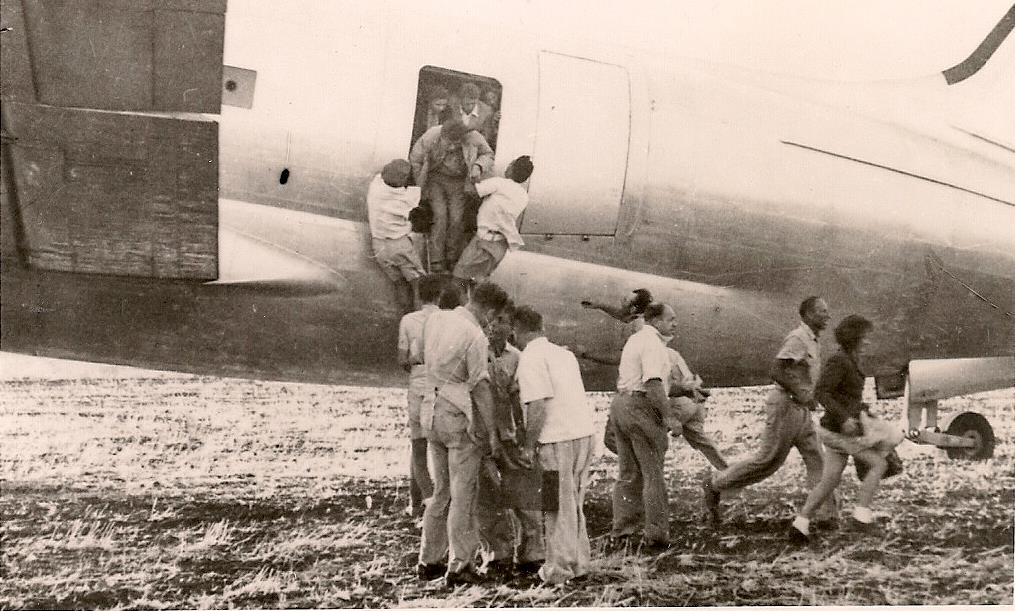 Jewish refugees boarding a propeller aircraft through a side door during an airlift, others waiting on the ground with belongings.
