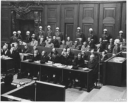 The defendants seated in two rows in the dock at the Nuremberg International Military Tribunal, with white-helmeted American military police standing behind them and defense counsel at desks in front.