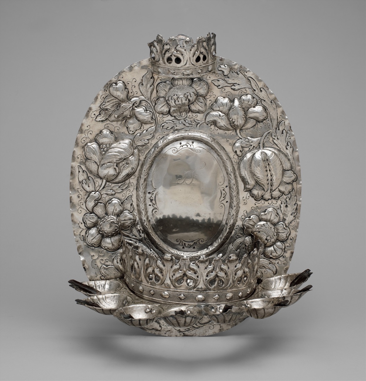 An ornate metal Hanukkah lamp with a decorated backplate and a row of eight oil lights