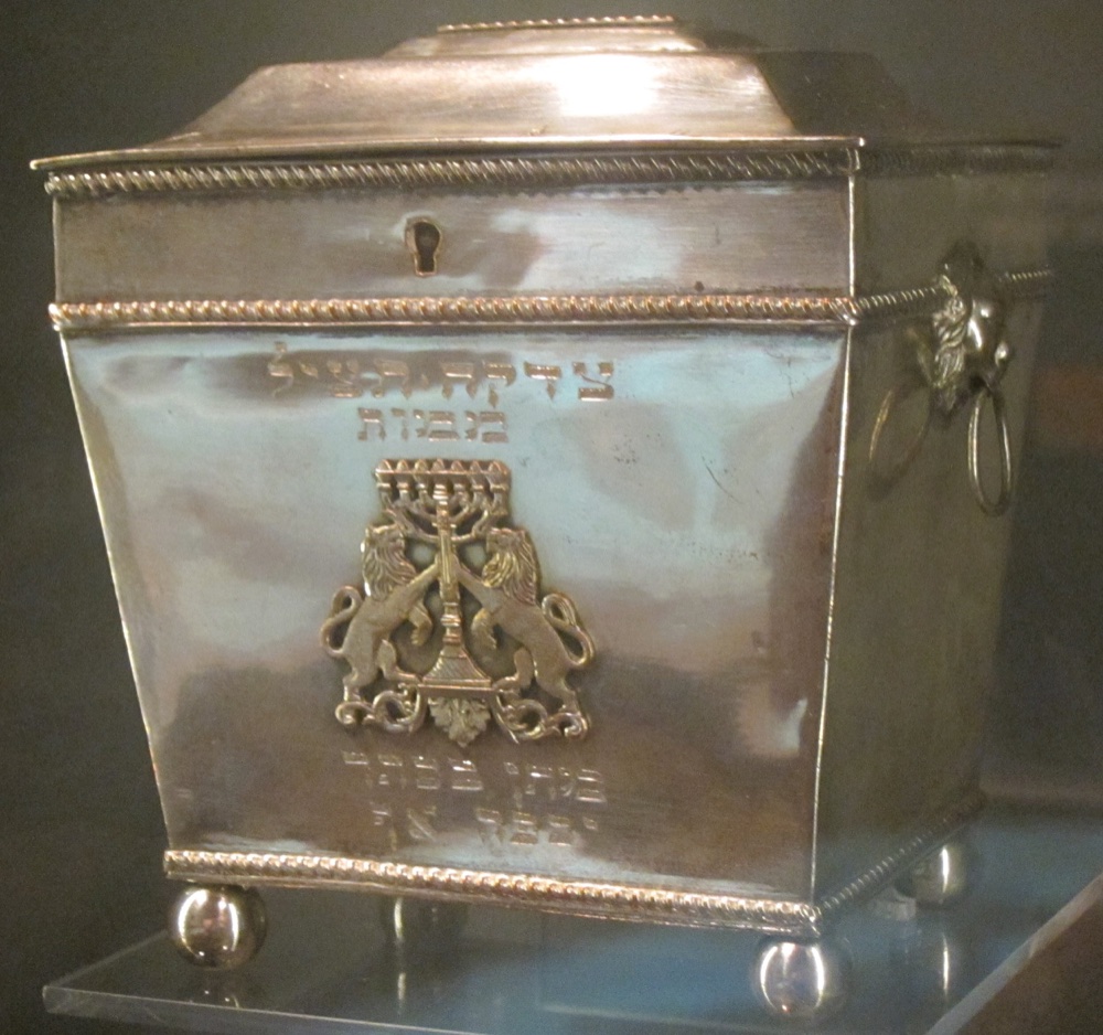 A silver tzedakah charity box with a menorah flanked by lions and a Hebrew inscription