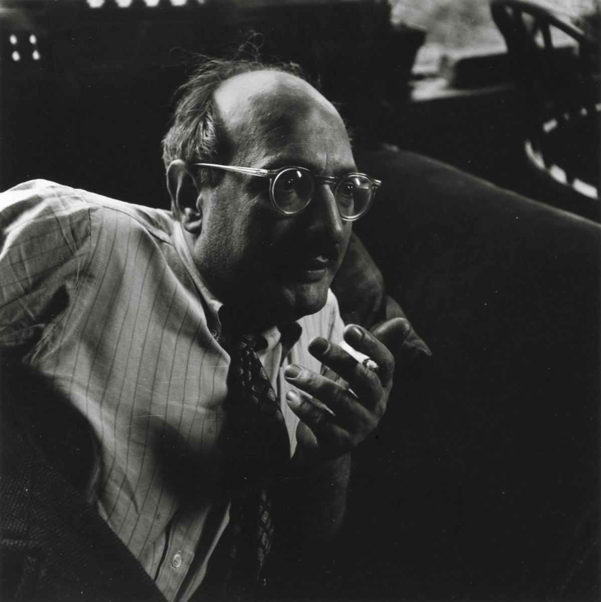 A black and white photograph of Mark Rothko in round glasses and a striped shirt, seated in shadow with a cigarette