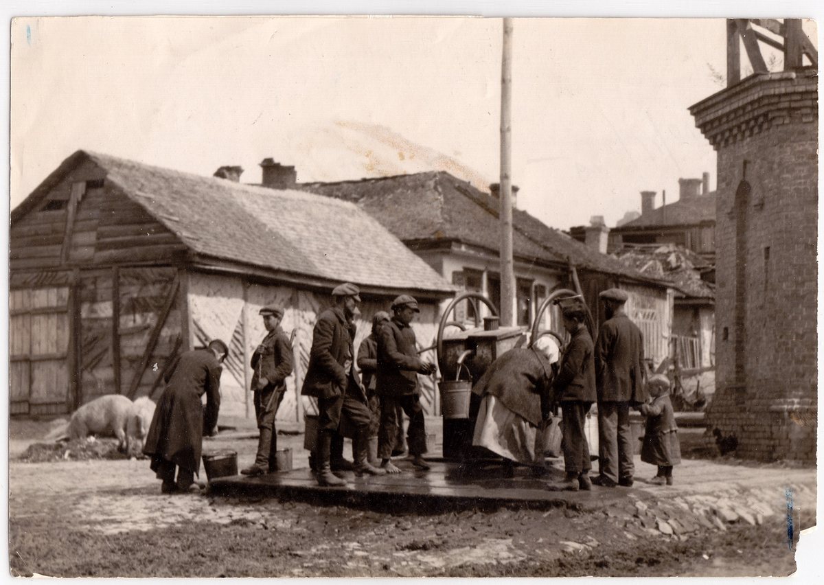 An archival photograph of townspeople drawing water at a communal well in a small Eastern European town, wooden houses behind them.