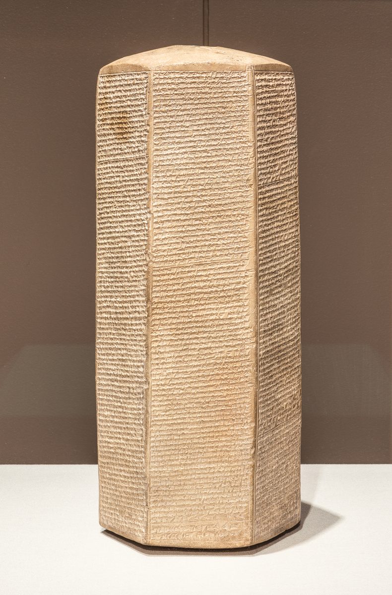 A six-sided baked-clay prism on a museum mount, every face covered in rows of cuneiform writing.