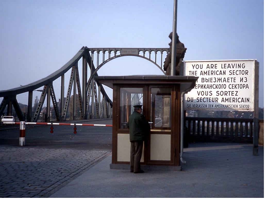 The West Berlin checkpoint at the Glienicke Bridge: a guard booth, a striped barrier, and a large sign reading &lsquo;You are leaving the American sector&rsquo; in English, Russian, French, and German, with the steel arch of the bridge behind.