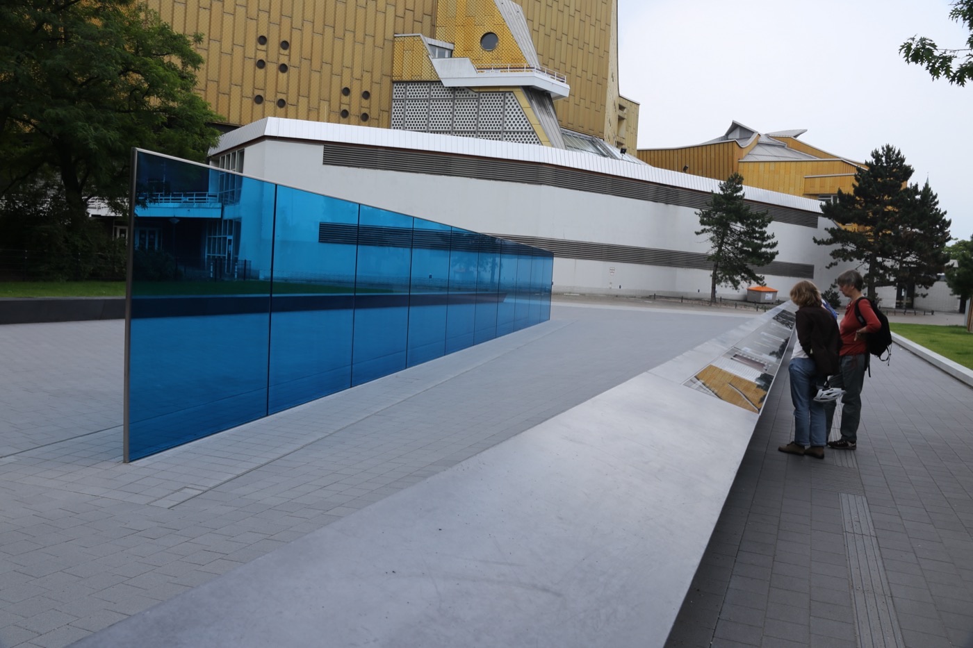 A long blue glass wall memorial on a paved plaza beside the golden Berlin Philharmonic building, with two visitors reading an information panel.