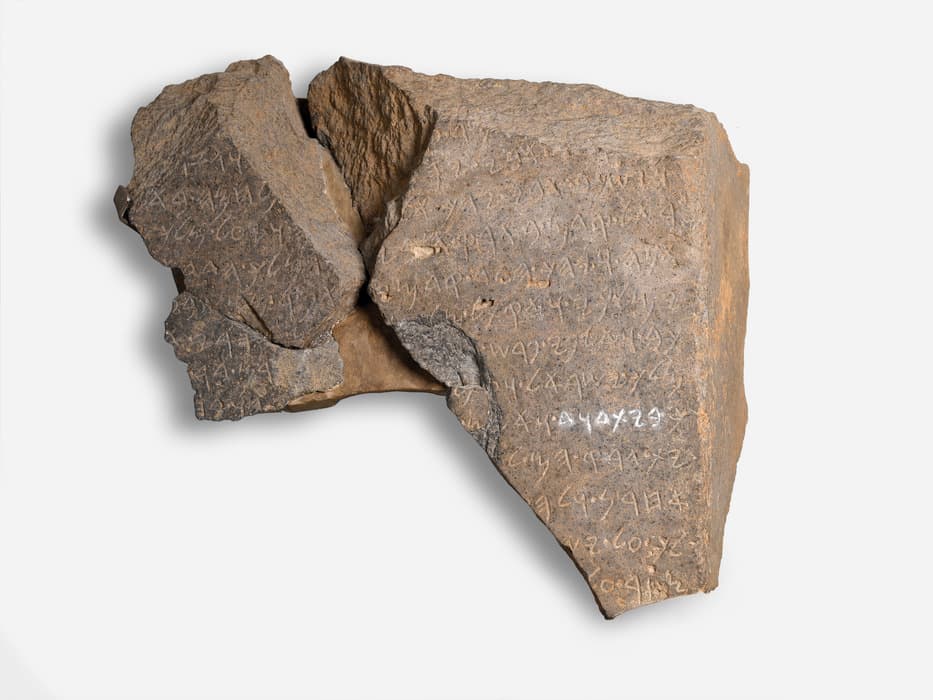 Several joined fragments of a dark basalt slab, the broken surface incised with rows of an early alphabetic script, one line highlighted in white.