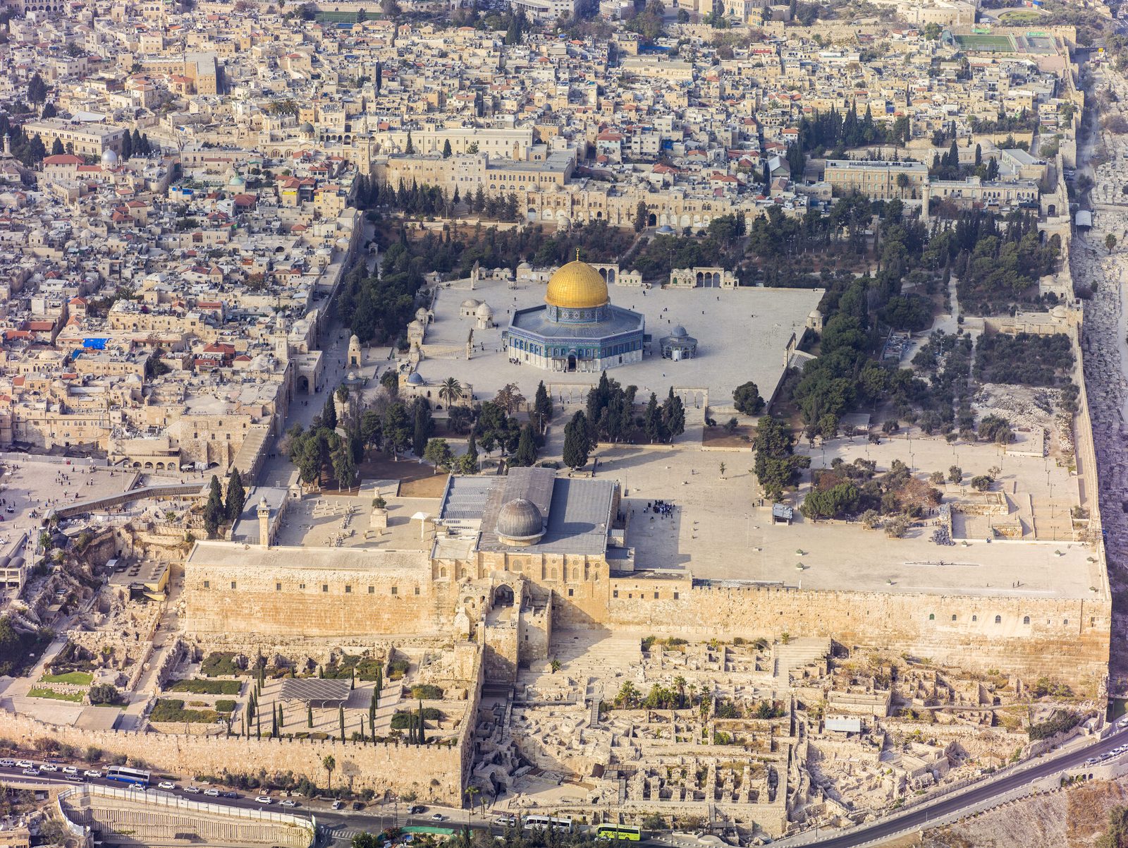 An aerial photograph of the walled Temple Mount platform in Jerusalem's Old City: a large paved plaza enclosed by ancient stone retaining walls, with the gold-domed Dome of the Rock near the center and the lead-domed al-Aqsa Mosque toward the lower edge, dense city around it.