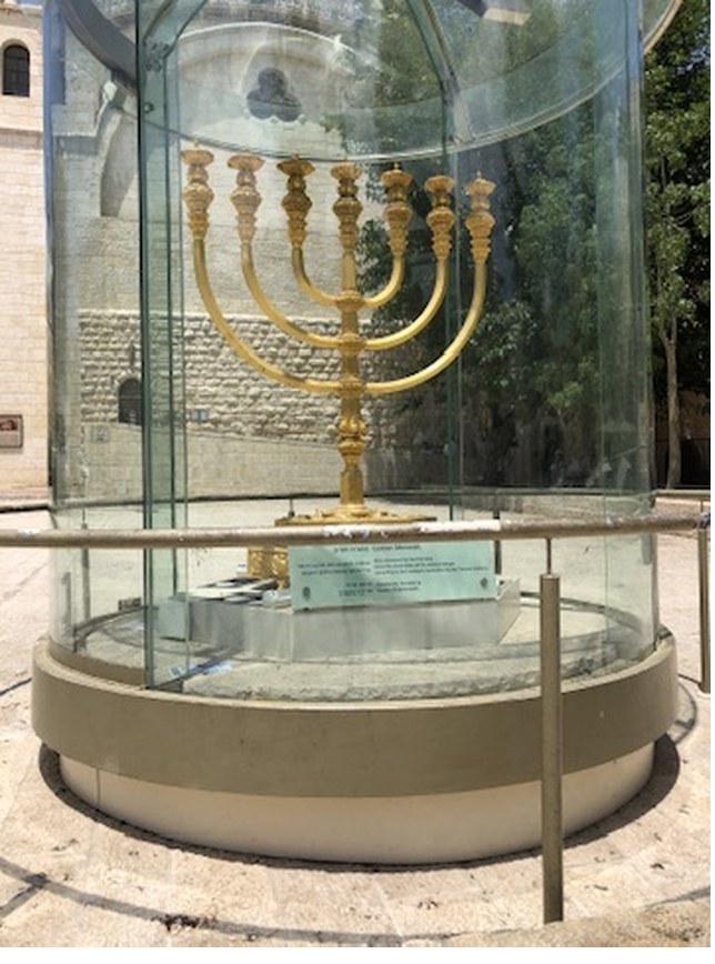 A large gold seven-branched menorah inside a cylindrical glass case in a Jerusalem street, with stone buildings behind it.