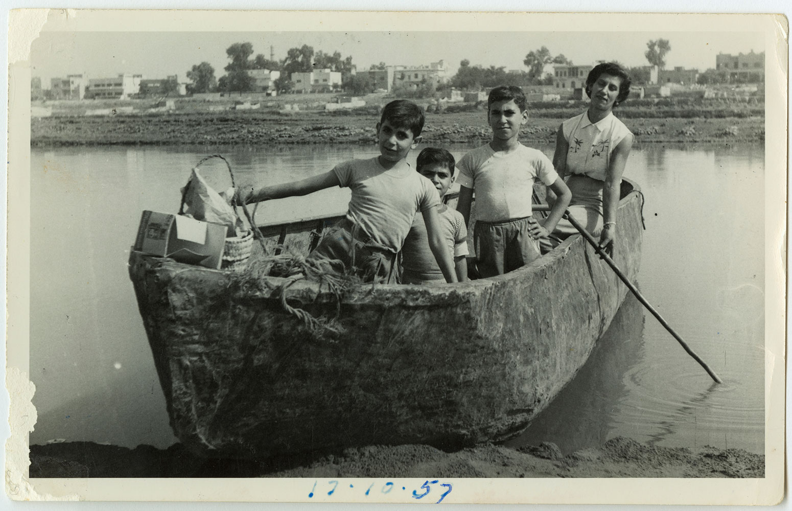 A black-and-white photograph of three boys and a young woman standing in a traditional reed-and-pitch boat on the Tigris River, with the low Baghdad skyline behind them. A picnic basket sits in the bow. The date 13-10-57 is written in blue ink at the bottom.
