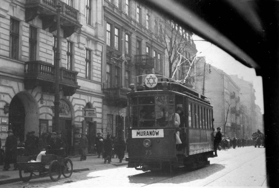A tram numbered 75 marked Muranów moves down a crowded Warsaw ghetto street, a Star of David mounted on its roof, with pedestrians, tenement buildings, and a handcart along the curb.