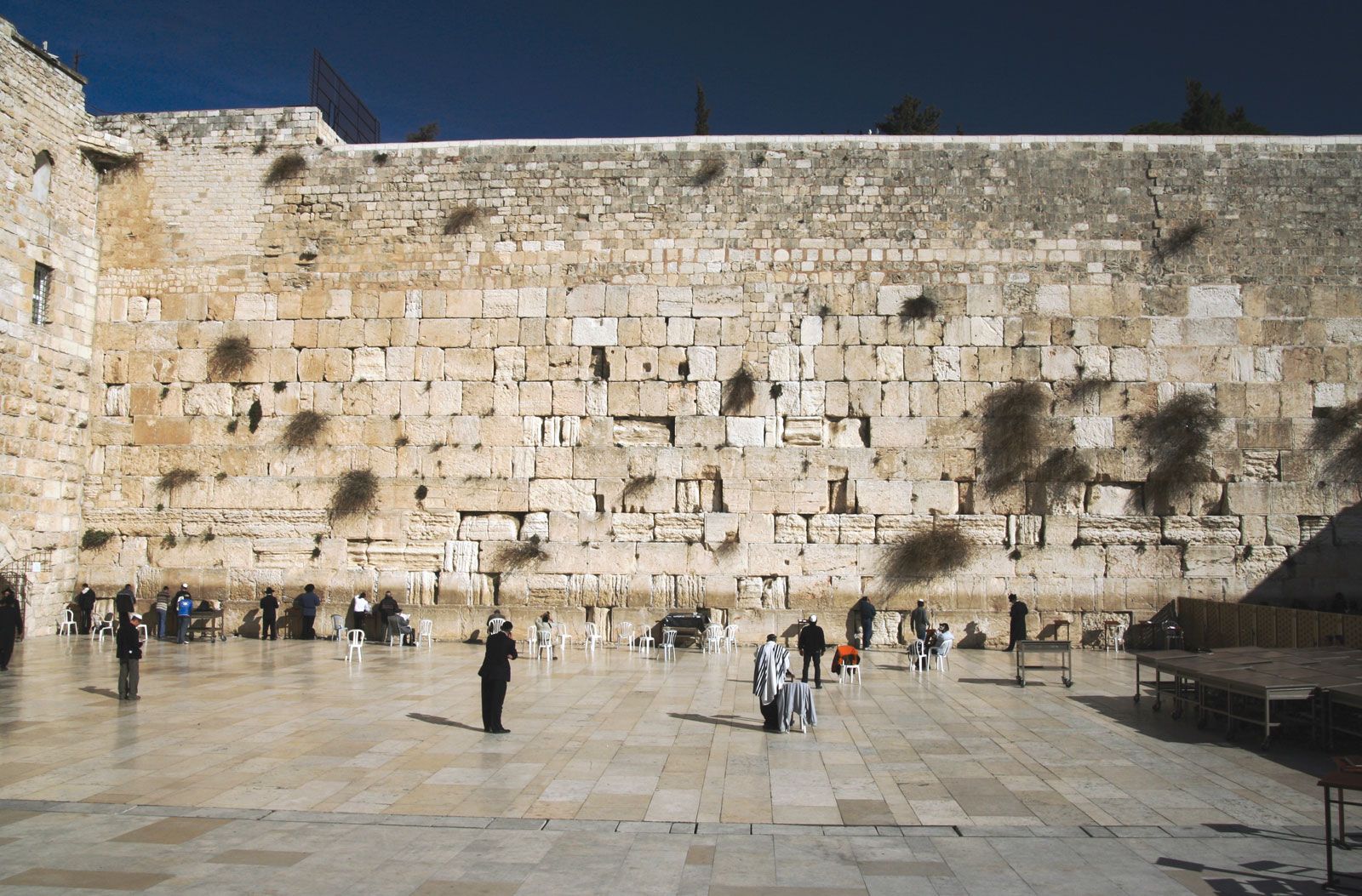The Western Wall and its open paved plaza in Jerusalem: a long, high wall of large weathered limestone blocks, with small clusters of people praying at its base, set against a deep blue sky.